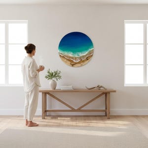 K&ouml;nnte beinhalten: Runde Wandkunst mit einer Strandlandschaft mit tiefblauem Ozean, t&uuml;rkisfarbenem Wasser und Sandstrand. Das Kunstwerk h&auml;ngt an einer wei&szlig;en Wand &uuml;ber einer Holzkonsolentisch. Eine Frau steht in der N&auml;he und h&auml;lt eine Tasse.