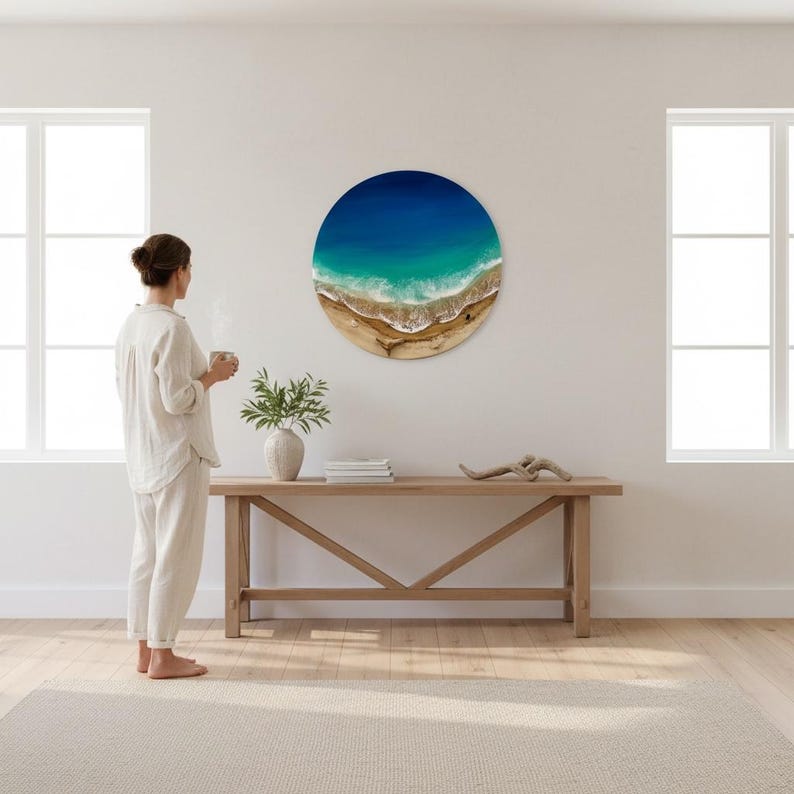 K&ouml;nnte beinhalten: Runde Wandkunst mit einer Strandlandschaft mit tiefblauem Ozean, t&uuml;rkisfarbenem Wasser und Sandstrand. Das Kunstwerk h&auml;ngt an einer wei&szlig;en Wand &uuml;ber einer Holzkonsolentisch. Eine Frau steht in der N&auml;he und h&auml;lt eine Tasse.