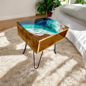 May include: A wooden end table with a clear epoxy resin top featuring a blue and green ocean wave design. The table has a lower shelf and black hairpin legs.