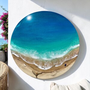 K&ouml;nnte beinhalten: Runde Wandkunst, die eine Strandszene mit einem hellblauen Himmel und t&uuml;rkisfarbenem Wasser darstellt. Das Kunstwerk zeigt einen sandigen Strand mit wei&szlig;en Wellen, einer Muschel und einer Walflosse. Der Durchmesser des Kunstwerks betr&auml;gt ca. 40 cm.