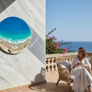 K&ouml;nnte beinhalten: Ein rundes Wandkunstwerk, das eine Strandszene mit blauem und t&uuml;rkisfarbenem Wasser, einem sandigen Ufer und wei&szlig;en Wellen darstellt. Das Kunstwerk ist an einer wei&szlig;en Marmorwand angebracht. Das Bild zeigt auch eine Person, die auf einer Terrasse mit Blick auf das Meer sitzt.