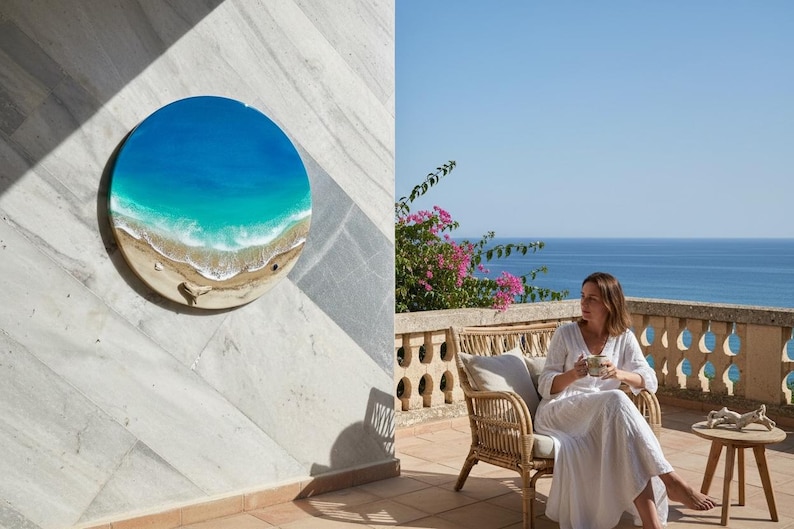 K&ouml;nnte beinhalten: Ein rundes Wandkunstwerk, das eine Strandszene mit blauem und t&uuml;rkisfarbenem Wasser, einem sandigen Ufer und wei&szlig;en Wellen darstellt. Das Kunstwerk ist an einer wei&szlig;en Marmorwand angebracht. Das Bild zeigt auch eine Person, die auf einer Terrasse mit Blick auf das Meer sitzt.