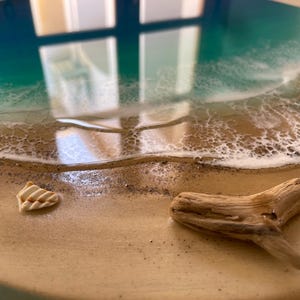 K&ouml;nnte beinhalten: Ein rundes Kunstwerk aus Kunstharz, das eine Strandszene darstellt. Es zeigt einen t&uuml;rkis-wei&szlig;en Ozean, einen Sandstrand, eine Muschel und ein St&uuml;ck Treibholz.