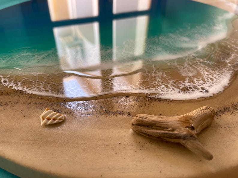 K&ouml;nnte beinhalten: Ein rundes Kunstwerk aus Kunstharz, das eine Strandszene darstellt. Es zeigt einen t&uuml;rkis-wei&szlig;en Ozean, einen Sandstrand, eine Muschel und ein St&uuml;ck Treibholz.