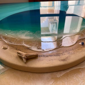 K&ouml;nnte beinhalten: Ein rundes Kunstwerk aus Harz mit Strandmotiv. Das Kunstwerk zeigt einen Farbverlauf aus Blau und T&uuml;rkis, der das Meer darstellt, mit einem sandigen Ufer und wei&szlig;en, sch&auml;umenden Wellen. Ein St&uuml;ck Treibholz und eine kleine Muschel sind im Sand eingebettet.