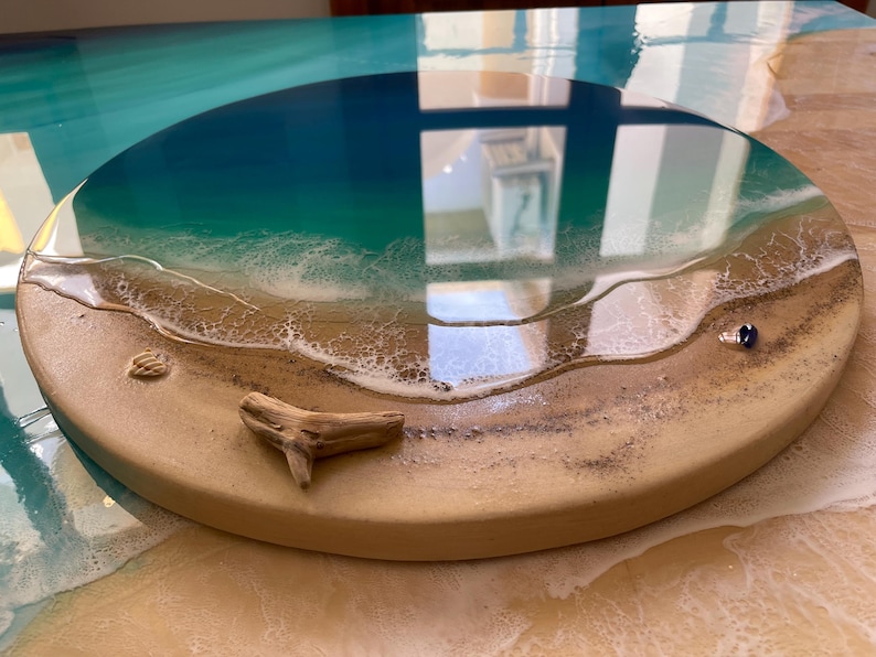 K&ouml;nnte beinhalten: Ein rundes Kunstwerk aus Harz mit Strandmotiv. Das Kunstwerk zeigt einen Farbverlauf aus Blau und T&uuml;rkis, der das Meer darstellt, mit einem sandigen Ufer und wei&szlig;en, sch&auml;umenden Wellen. Ein St&uuml;ck Treibholz und eine kleine Muschel sind im Sand eingebettet.