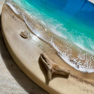 K&ouml;nnte beinhalten: Ovale Kunstharz-Kunstwerk, das eine Strandszene darstellt. Das Kunstwerk zeigt einen sandigen Strand mit kleinen Muscheln und einem St&uuml;ck Treibholz. Das Meer ist in Blau- und T&uuml;rkist&ouml;nen gehalten, mit wei&szlig;en, sch&auml;umenden Wellen. Das St&uuml;ck befindet sich auf einer hellen Holzbasis.