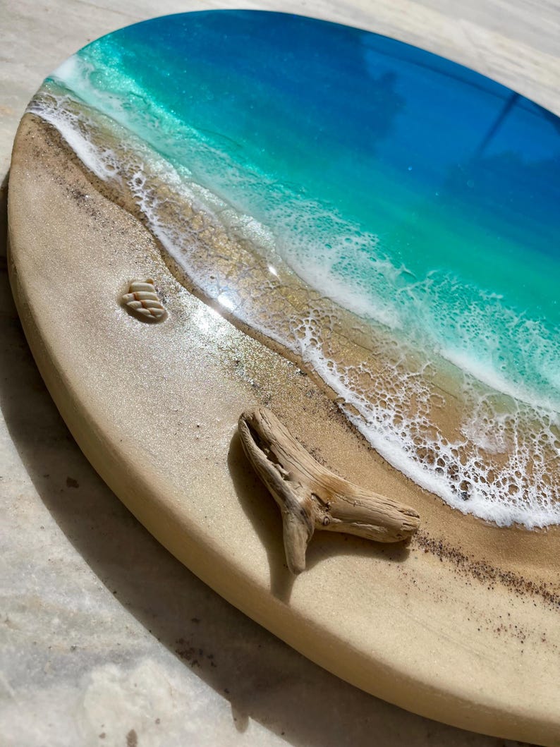 K&ouml;nnte beinhalten: Ovale Kunstharz-Kunstwerk, das eine Strandszene darstellt. Das Kunstwerk zeigt einen sandigen Strand mit kleinen Muscheln und einem St&uuml;ck Treibholz. Das Meer ist in Blau- und T&uuml;rkist&ouml;nen gehalten, mit wei&szlig;en, sch&auml;umenden Wellen. Das St&uuml;ck befindet sich auf einer hellen Holzbasis.