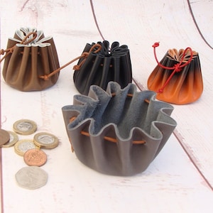 May include: Four small leather pouches in brown, black, grey, and orange with drawstring closures. The pouches are sitting on a white wooden surface with some coins scattered around them.