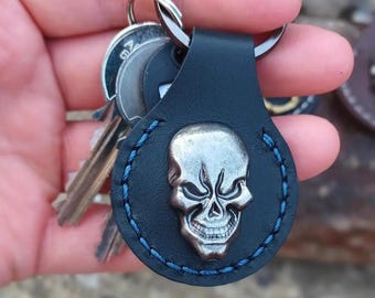May include: A black leather keychain with a silver skull charm. The keychain has blue stitching around the edges.