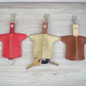 May include: Three leather keychains in the shape of a t-shirt. The keychains are red, gold, and brown, with contrasting stitching. Each keychain has a silver clasp and a leather strap.