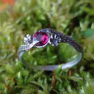 Könnte beinhalten: Ein silberner Ring mit einem rosafarbenen Edelstein und floralen Designs. Der Ring liegt auf einem Bett aus grünem Moos.