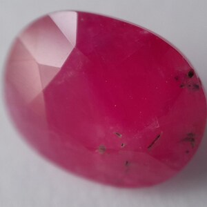 May include: A pink oval-shaped gemstone with a faceted cut. The gemstone has some black inclusions.
