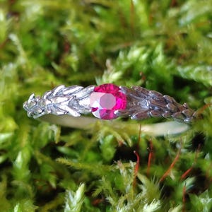 Puede incluir: Un anillo de plata con una piedra preciosa roja brillante engastada en el centro. La banda del anillo está decorada con un diseño de hojas.