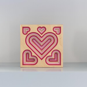 Peut inclure: Une œuvre d'art murale en forme de cœur en bois avec des cœurs roses et rouges superposés. L'œuvre est carrée, avec un cadre en bois naturel et une série de cœurs imbriqués. Des accents décoratifs sont de chaque côté.