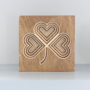 Peut inclure: Décor mural en bois en forme de trèfle. Le motif de trèfle à trois feuilles est sculpté dans un bloc de bois carré. Les feuilles du trèfle ont la forme de cœurs. Le bois a une teinte marron chaud et naturelle. Article décoratif.