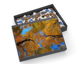 May include: A jigsaw puzzle featuring a vibrant autumn scene of trees with golden and orange leaves against a bright blue sky. The puzzle is in a black box, with some pieces visible.