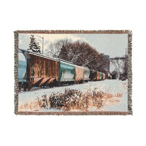 May include: Woven throw blanket depicting a winter scene with a train of boxcars in the snow. The blanket has a fringed edge and shows a snowy landscape with trees and a building in the background.