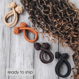 Può includere: Accessori per capelli in vari colori, tra cui beige, arancione, marrone e nero, disposti su una superficie di legno. Gli accessori sono ad anello e annodati, con una sezione di capelli ricci marroni sullo sfondo. Il testo "ready to ship" è visibile.