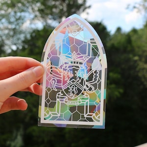 May include: A white sticker with a stained glass design featuring a coat of arms with a crown, animals, and a butterfly. The text "AMANS" is at the bottom of the sticker.