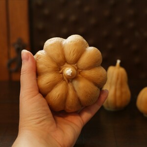 May include: A set of three brown pumpkin-shaped candles. The largest candle is being held in a hand. The candles are made of wax and have a smooth, textured surface.
