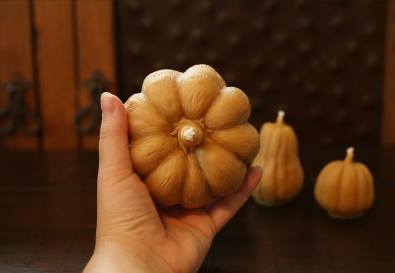 May include: A set of three brown pumpkin-shaped candles. The largest candle is being held in a hand. The candles are made of wax and have a smooth, textured surface.