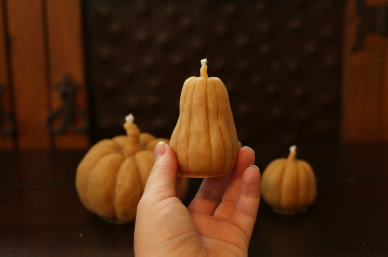 May include: Three small, brown, pumpkin-shaped candles. The candle in the center is being held by a hand.