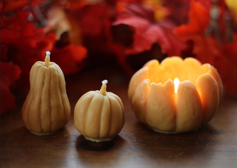 May include: Three decorative pumpkin-shaped candles, two are off and one is lit, on a wooden surface with a blurred background of red fall leaves.