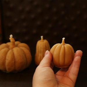May include: Three small, hand-crafted, pumpkin-shaped candles. The candles are made of a light brown wax and have a white wick. The candles are arranged on a dark brown wooden surface.