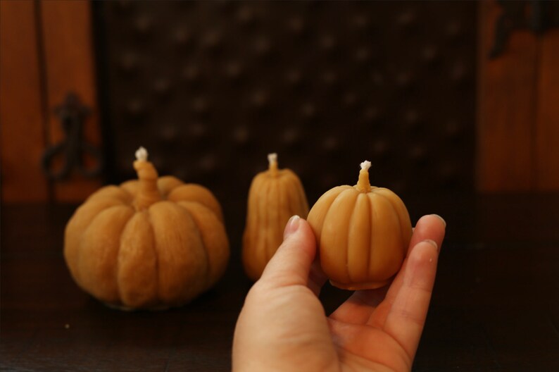 May include: Three small, hand-crafted, pumpkin-shaped candles. The candles are made of a light brown wax and have a white wick. The candles are arranged on a dark brown wooden surface.