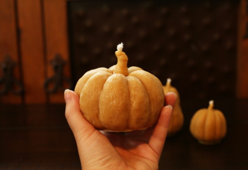 May include: A hand holds a brown, pumpkin-shaped candle with a white wick. Two smaller, similar candles are in the background.
