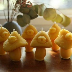 May include: Six yellow beeswax candles shaped like mushrooms with a single eye on each cap. The candles are arranged on a wooden surface.