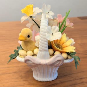 May include: A decorative Easter basket with a yellow duckling, white and yellow flowers, and pink accents. The white basket has a handle and is filled with faux flowers and decorative beads. A charming spring-themed decoration.