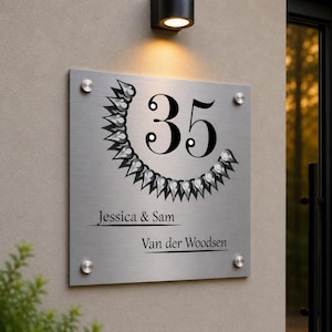May include: A rectangular brushed metal address sign displaying the number 35 in black, encircled by a black decorative pattern. The sign also features the names "Jessica & Sam" and "Van der Woodsen". It is mounted on a beige wall with a light fixture above.