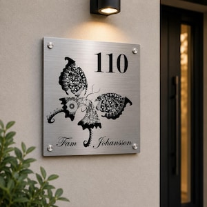 May include: A rectangular brushed metal address plaque displaying the number "110" in black. A detailed butterfly design is etched below the number. The name "Fam Johansson" is inscribed at the bottom. The plaque is mounted on a light-coloured wall.