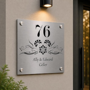 May include: A square, brushed metal house number sign displaying the number 76 in black. Below the number is a floral design and the names "Ally & Edward Geller" in black. The sign is secured with four silver screws.