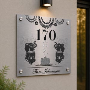 May include: A brushed metal house number sign displaying the number 170 in black. The sign features two elephant illustrations, a lotus flower, and the name "Fam Johansson" in script. Decorative floral patterns are at the top.