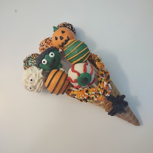 May include: A waffle cone filled with Halloween-themed cake pops. The cake pops are decorated with green, orange, white, and black frosting and sprinkles. The cake pops are shaped like a pumpkin, a monster, a mummy, and an eyeball.