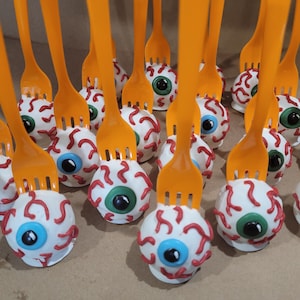 May include: A close-up of a group of eyeball cake pops on orange plastic forks. The cake pops are decorated with white frosting, red veins, and blue and green eyeballs.