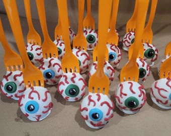 Könnte beinhalten: Nahaufnahme einer Gruppe von Augen-Cake-Pops auf orangefarbenen Plastikgabeln. Die Cake-Pops sind mit weißem Frosting, roten Adern und blauen und grünen Augen verziert.