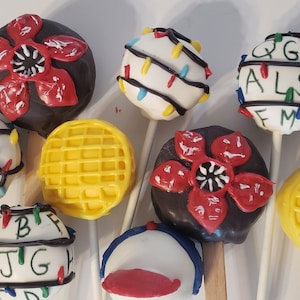May include: A collection of eight Stranger Things themed cake pops. The cake pops are decorated with various designs, including the Upside Down, Christmas lights, and the iconic Eggo waffle.