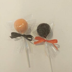 May include: Two cake pops, one orange and one black, are wrapped in clear cellophane and tied with ribbon. The orange cake pop has a black ribbon and the black cake pop has an orange ribbon.