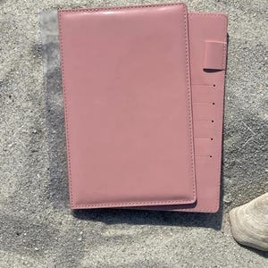 May include: A pink leather notebook with a matching pen holder and several card slots. The notebook is rectangular and has a smooth, glossy finish. The notebook is lying on a sandy surface.