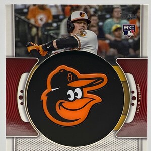 May include: A baseball card featuring a commemorative patch of the Baltimore Orioles logo. The logo is a black and orange cartoon bird with a baseball cap. The card also features a photo of a baseball player in a white uniform with the Orioles logo on the sleeve. The text "BAL" and "ADLEY RUTSCHMAN" are printed on the card.