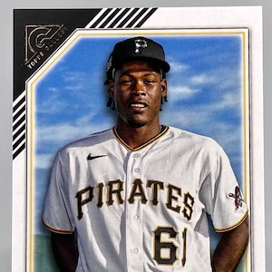 May include: A baseball card featuring a baseball player wearing a white jersey with the word "PIRATES" and the number "61" on the front. The player is wearing a black cap with a white "P" on the front. The card has a gold and black border with the words "TOPS GALLERY" and "PRINTER PROOF" on it. The player's name, "ONEIL CRUZ", is printed at the bottom of the card.