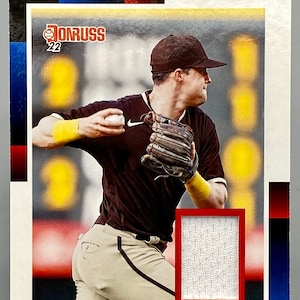 May include: A baseball card featuring Jake Cronenworth of the San Diego Padres. The card is red, white, and blue with a photo of Cronenworth in a brown uniform throwing a baseball. The card also includes a white jersey patch.