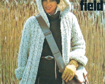 May include: A white knitted hooded cardigan with a brown leather strap and a beige bag. The cardigan is worn over a black turtleneck and black pants. The text "Taking the field" is in the upper right corner of the image.