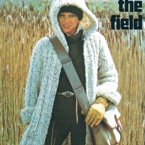 May include: A white knitted hooded cardigan with a brown leather strap and a beige bag. The cardigan is worn over a black turtleneck and black pants. The text "Taking the field" is in the upper right corner of the image.