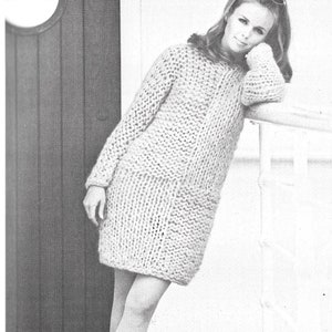 May include: A woman wearing a long, light-colored knitted sweater dress. She is standing next to a white railing and has her arm resting on it. She is wearing sunglasses and has her hair pulled back.