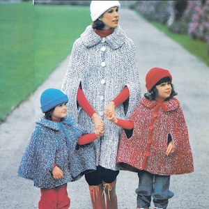 May include: Three people wearing knitted capes and hats. The adult is wearing a white hat and a gray cape with white buttons. The two children are wearing blue and red capes with matching hats.